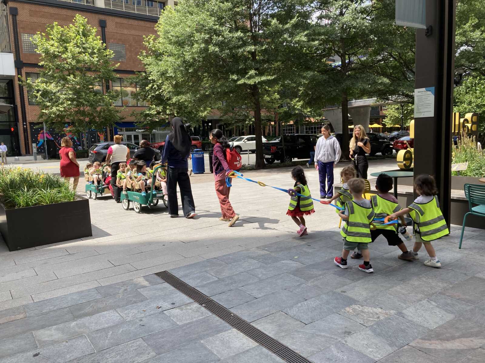 A kid train in Ballston (courtesy Gale Harris) | ARLnow Group of young children in neon vests ride in green wagon carts, pulled by adults in an urban plaza.