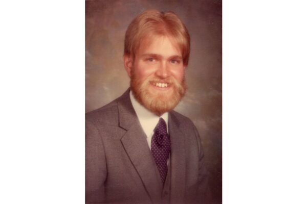 Portrait of a smiling man with blond hair and beard, wearing a gray suit and purple tie.