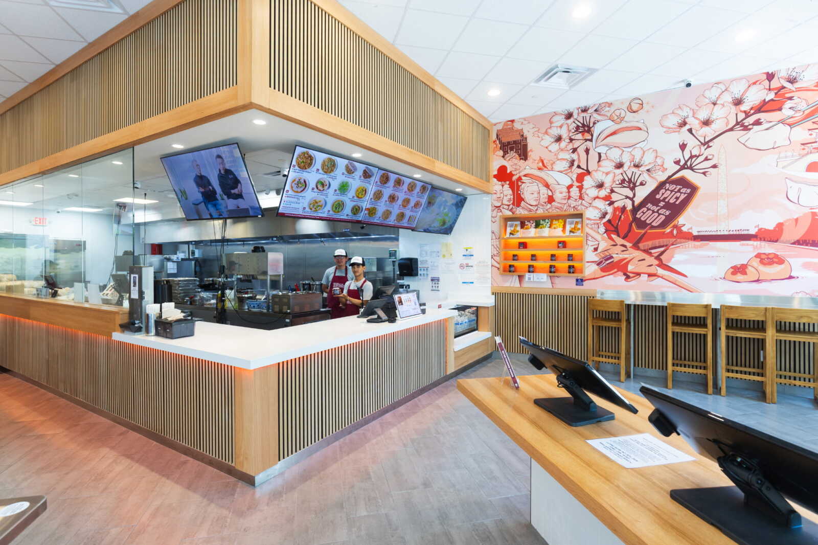 Restaurant counter area with two staff behind the white prep counter and digital menu boards overhead, kitchen visible in back, and a mural wall to the right.