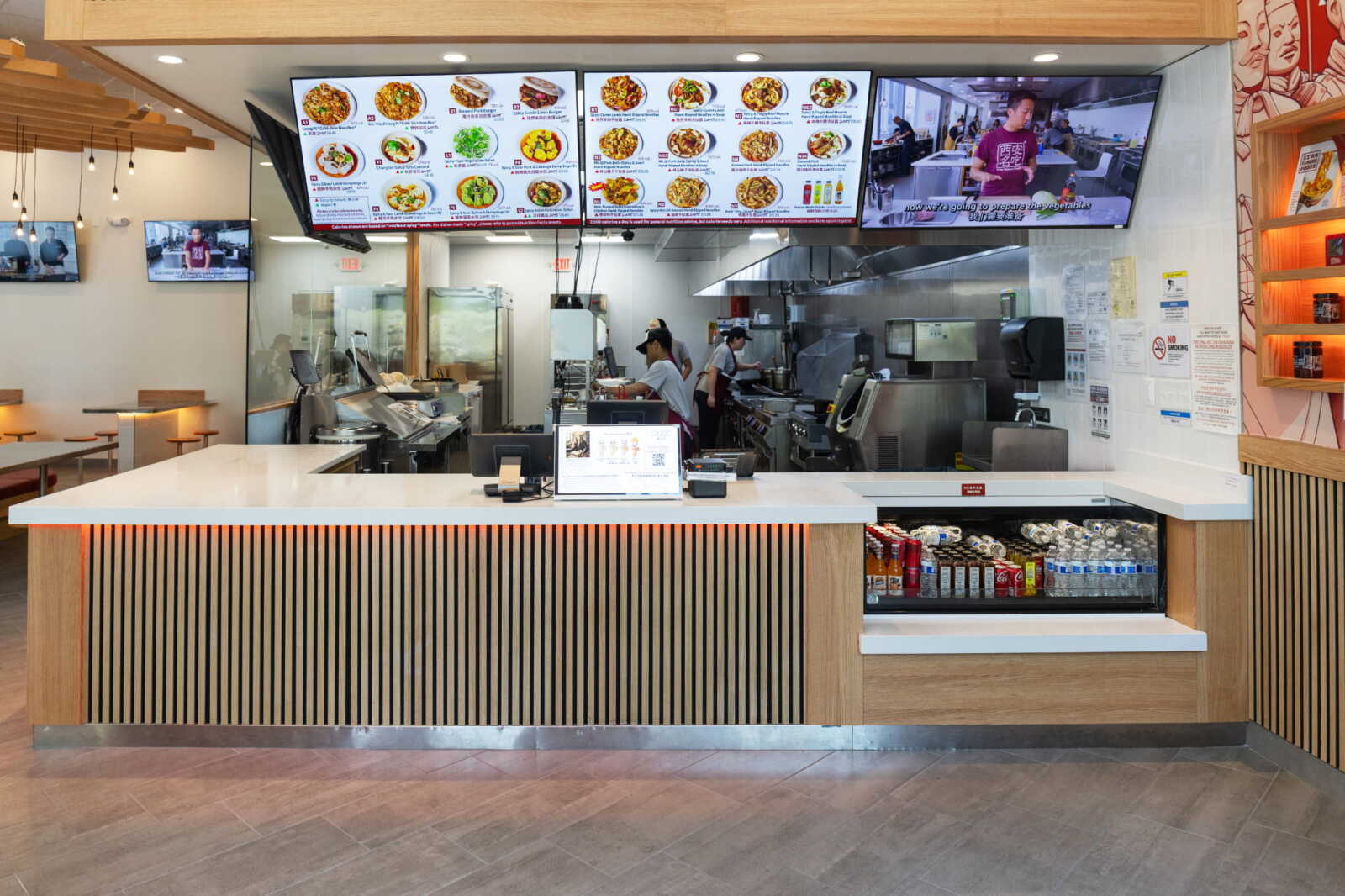 Front counter with vertical wood slats and illuminated orange edge, open kitchen in the background and digital menu boards overhead.