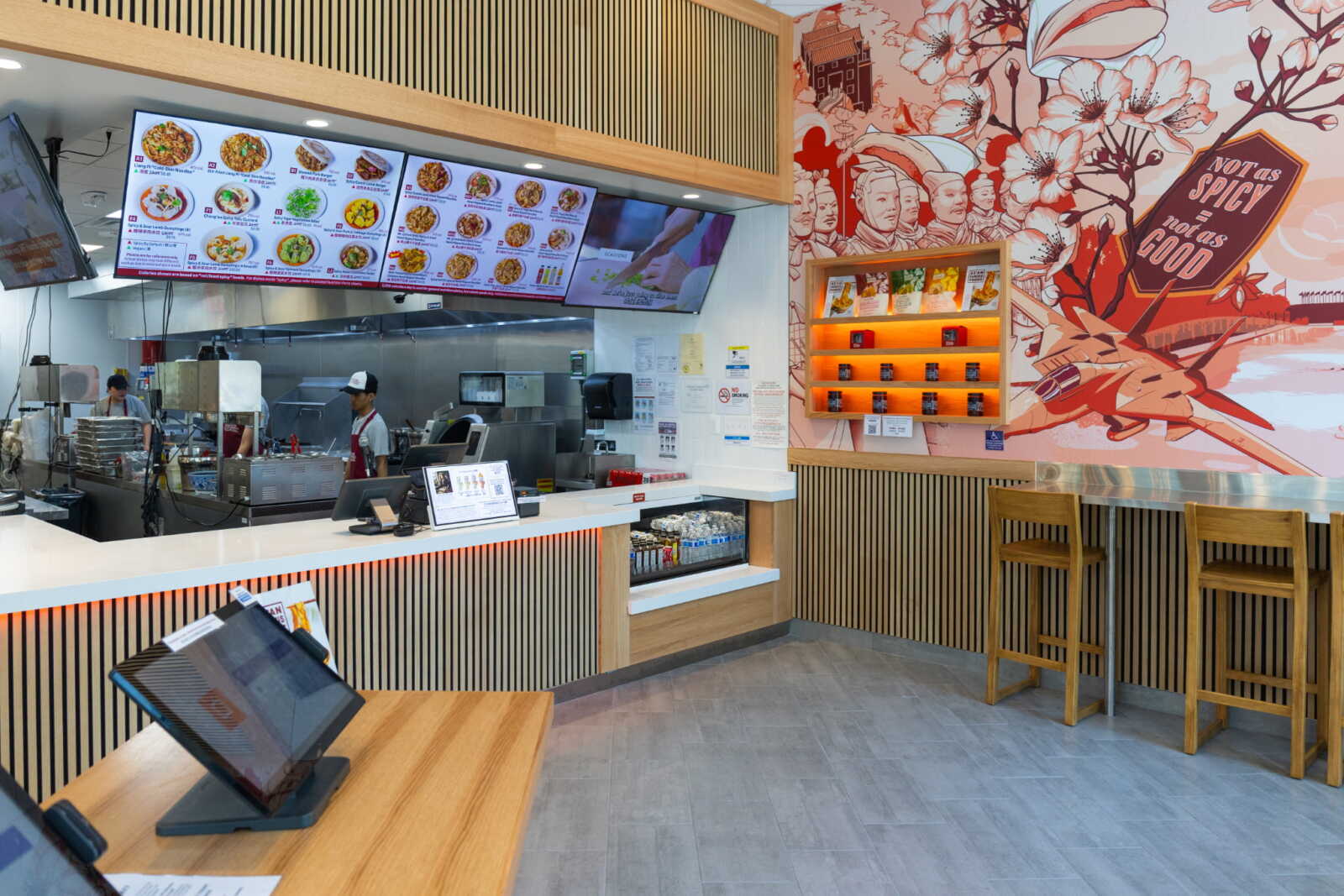 Restaurant counter with digital menu boards overhead and open kitchen in back, stools along the bar, and a colorful mural on the wall.