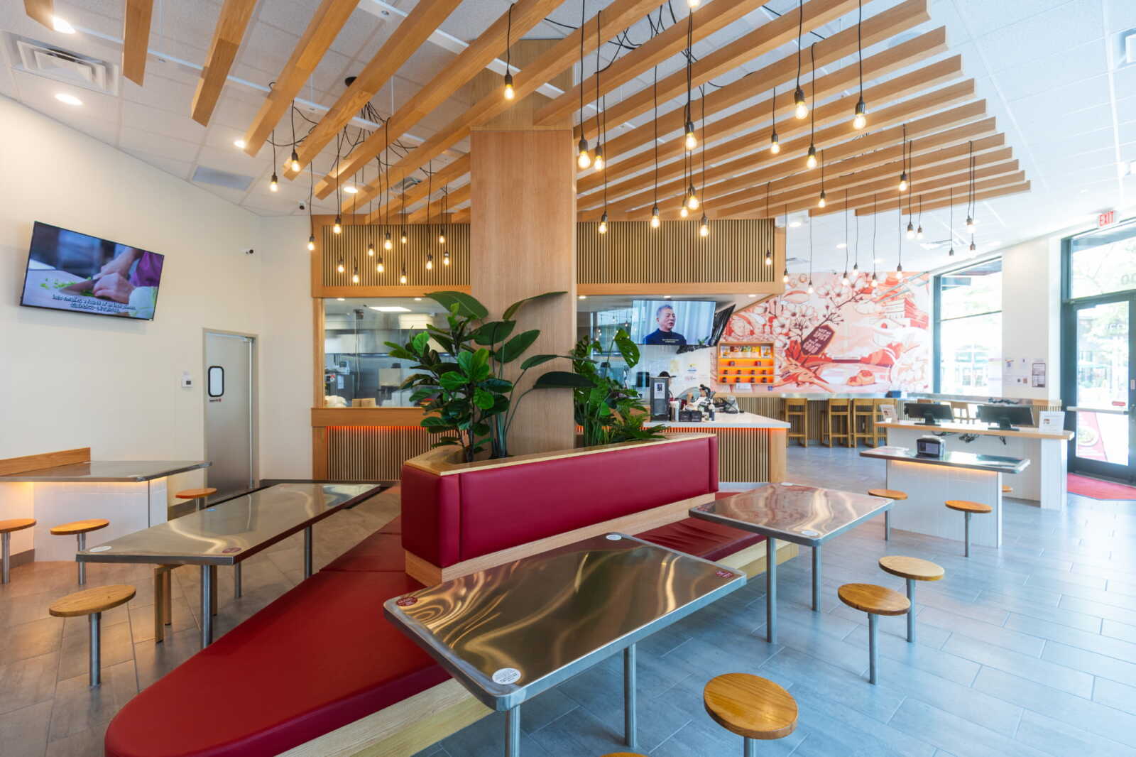 Bright modern cafe interior with wooden slat ceiling, hanging bulbs, a red booth, metal tables, and potted plants.