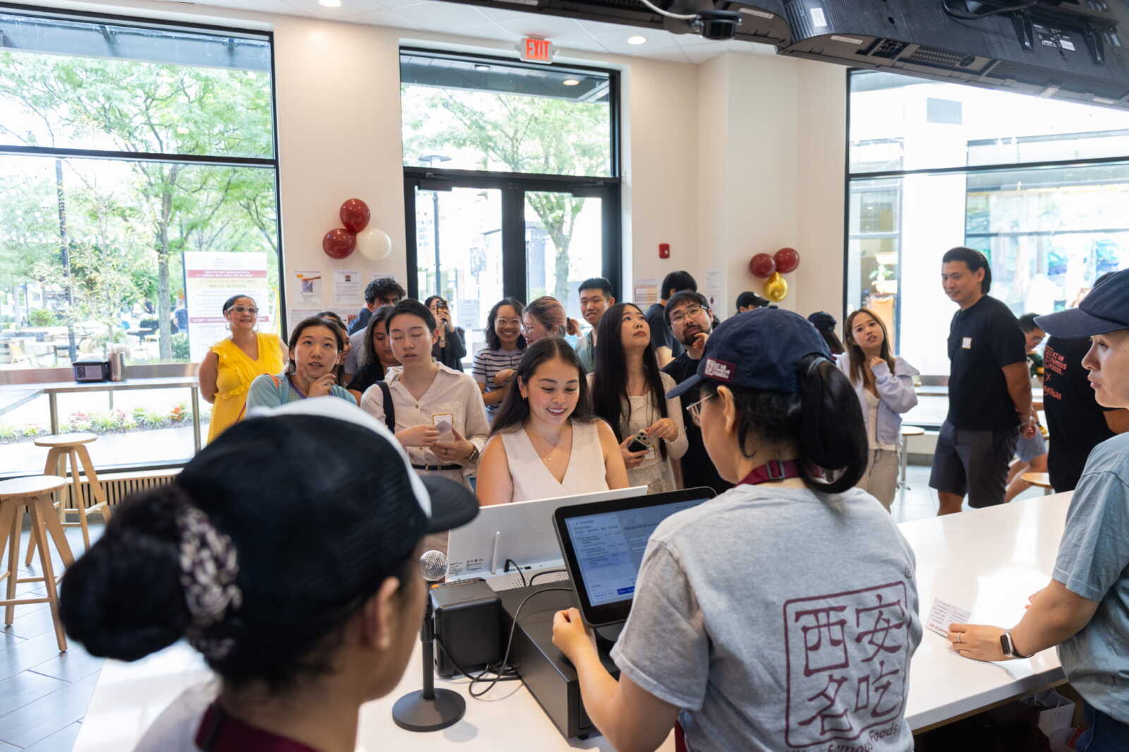 Crowd of people waiting in line at a bright cafe counter, staff using tablets at the front. balloons on the wall suggest a celebration or event.