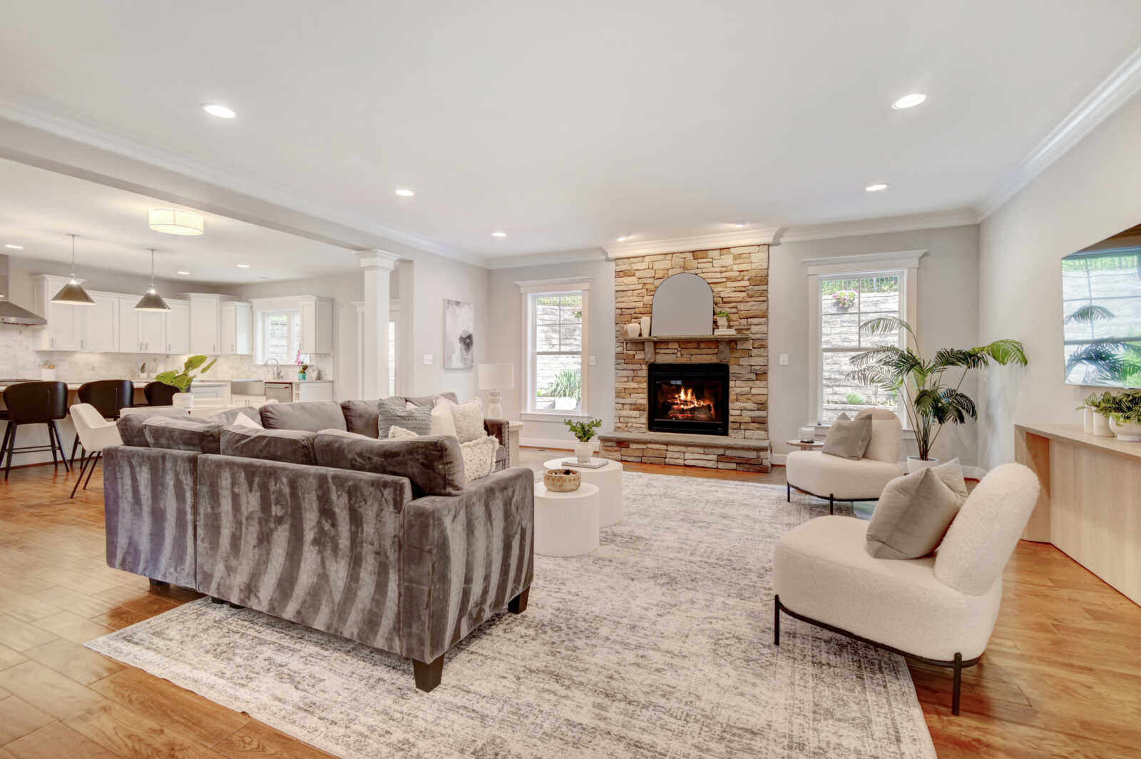 Open living room with a stone fireplace, gray sofa, white chairs, and a large area rug in a bright, airy space.