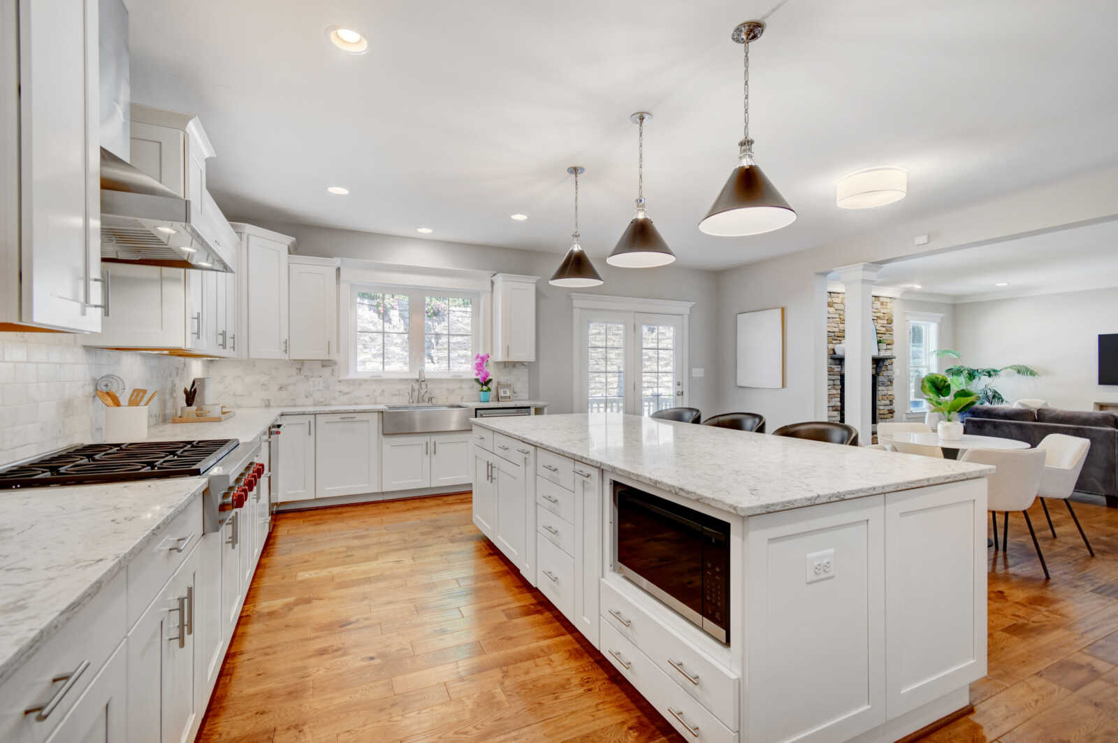Bright white kitchen with marble countertops, island, stainless steel stove, and hardwood floors; open to living area behind. - (informative)