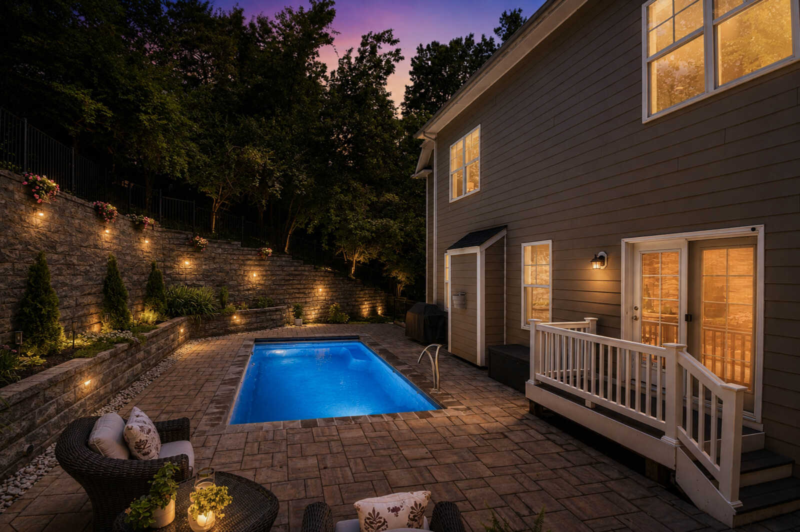 Backyard at dusk with a rectangular, blue-lit pool, a stone retaining wall with planters, and a beige house with lit windows.