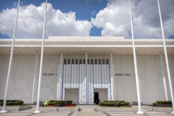 White modern building facade with construction tarp over the entrance; the space is labeled a performing arts venue, blue sky with clouds, flagpoles in foreground, and a worker in high-visibility gear nearby.
