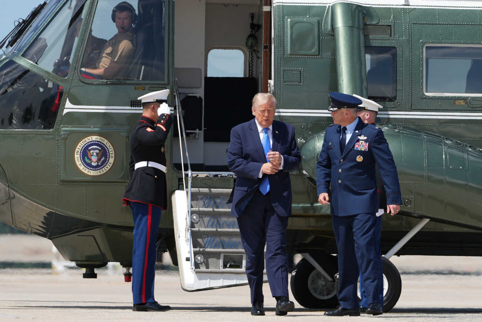 A man in a dark suit steps off a green presidential helicopter, flanked by two uniformed officers.