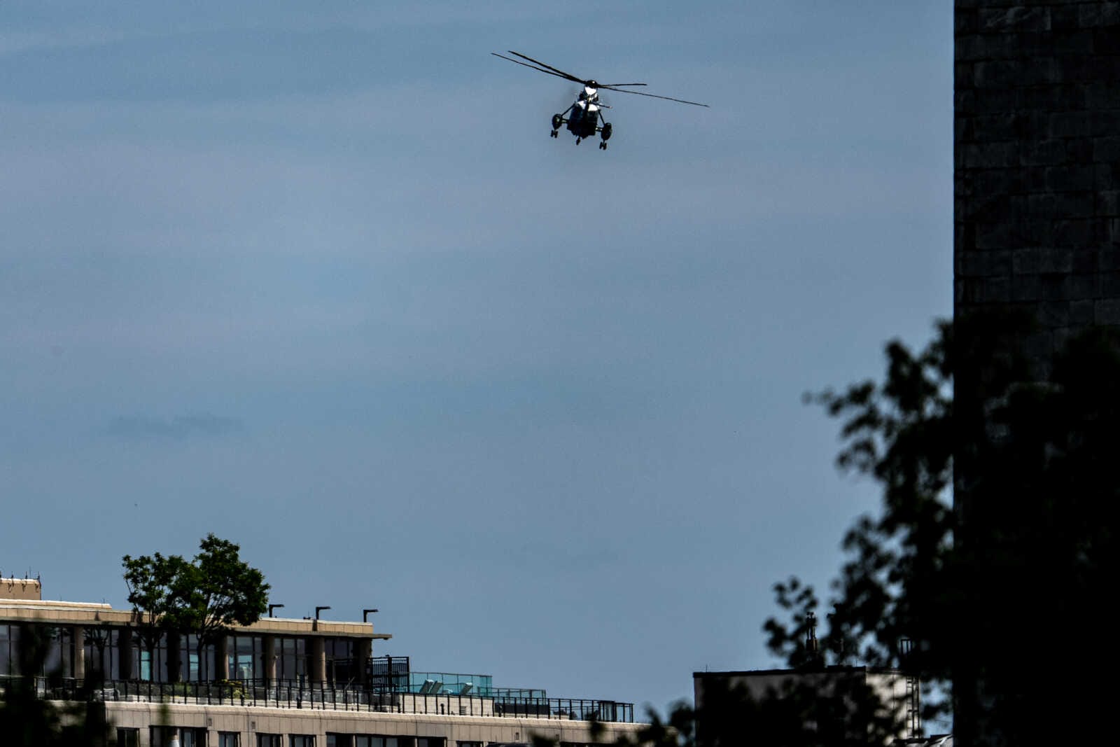 A helicopter hovering in the blue sky above a city block with trees on the left and a dark building edge on the right.