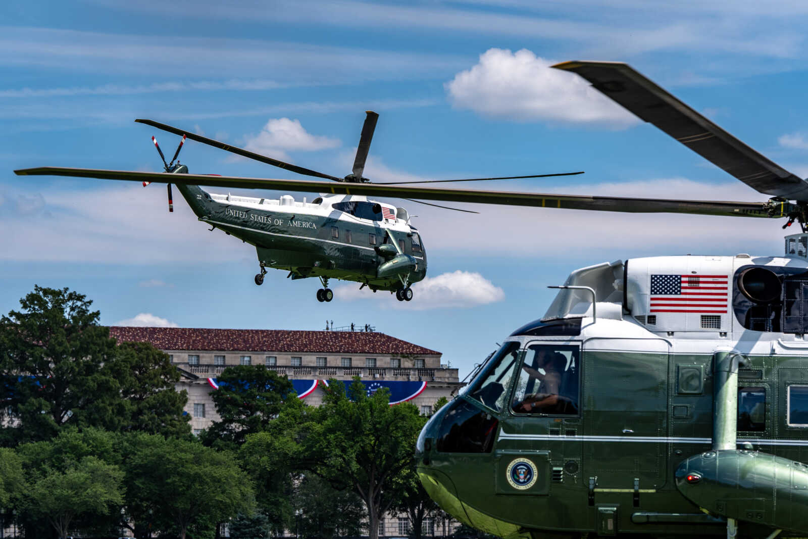 Green US military helicopter hovering above a government building, with another helicopter on the right and patriotic bunting in the background.