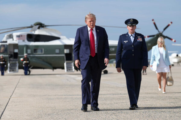 Donald Trump walking on an airfield beside a uniformed officer, with a large helicopter in the background.