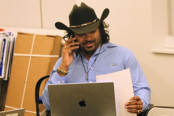 Man in a cowboy hat on a phone call at a desk, with a MacBook and papers in hand.