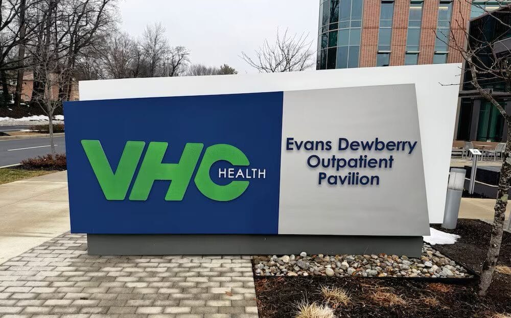 Outdoors sign for VCH Health: large blue panel with green VCH letters and the words 'Evans Dewberry Outpatient Pavilion' on a gray panel nearby
