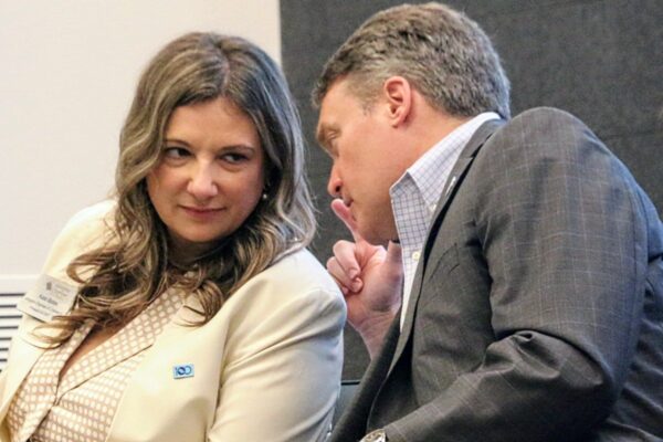 Two professionals in a conversation: a woman in a cream blazer and polka-dot blouse listens as a man in a gray suit leans in to speak to her at a conference edge.