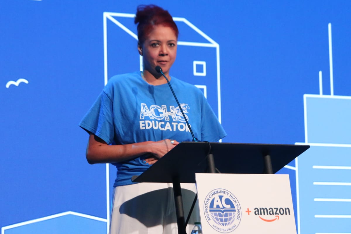 Woman in a blue ACHS Educator T-shirt gives a speech at a podium with event signage showing logos for Arlington Community High School and Amazon in the background.