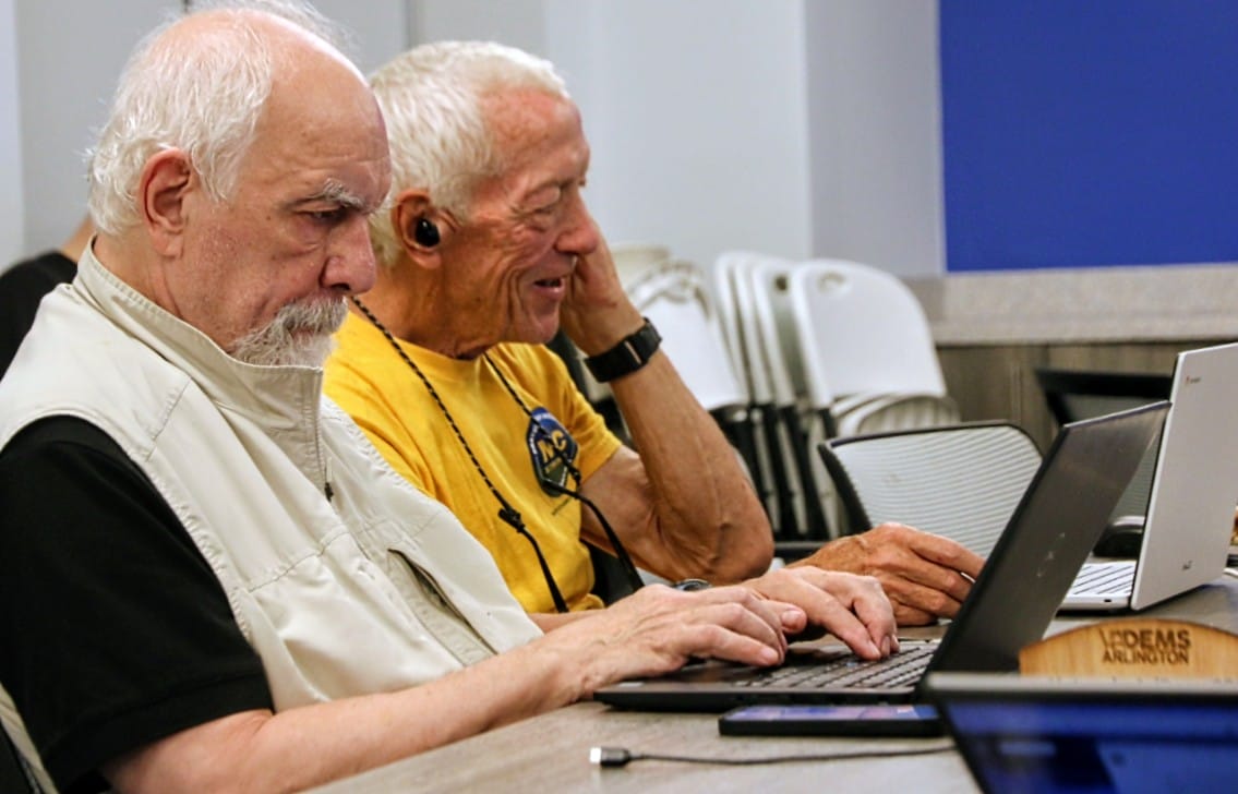 Two elderly men sit at a table, typing on laptops during a computer class or workshop in a community space.