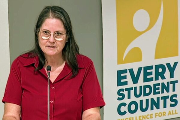 Woman with shoulder-length dark hair and glasses speaks at a microphone, wearing a red blouse, beside a banner that says 'EVERY STUDENT COUNTS'.