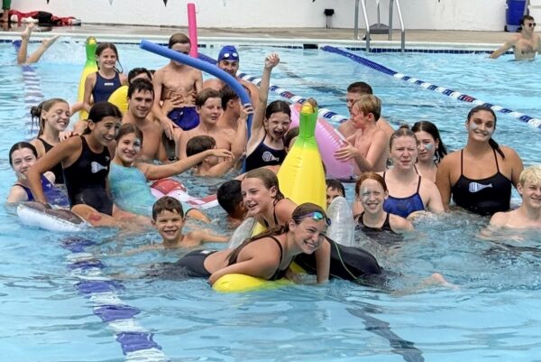 Group of children in a swimming pool with colorful inflatables and pool noodles, posing and smiling.