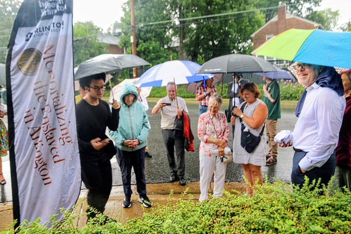 Group of people standing in the rain under umbrellas at an outdoor event, with a tall banner on the left side.