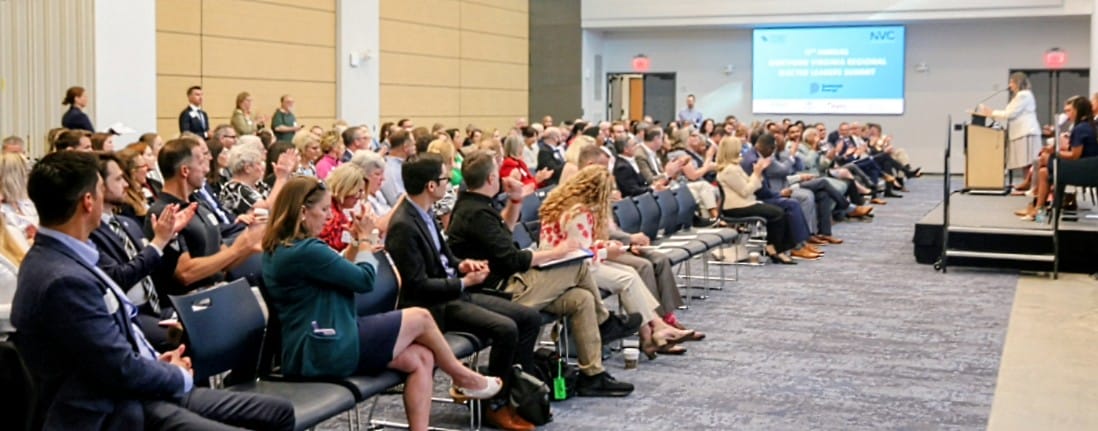 Large conference audience seated in rows, clapping as speakers present on a stage with a podium and screen in the background.