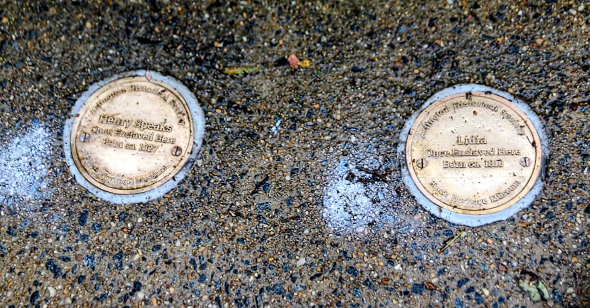 Two round commemorative plaques set in a wet sidewalk; one reads Henry Speaks and the other Lidia, with birth years around 1810.