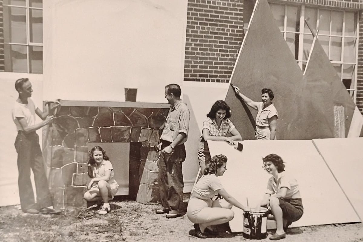Six people, adults and children, work together outdoors on a construction project against a brick building, assembling a faux stone wall with a doorway and large triangular shapes nearby.