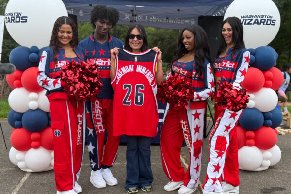 Group of five people posing with a red basketball jersey #26 at a Wizards-themed event, cheerleaders flank the center holder under balloon arches.