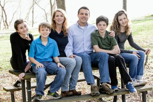 Family of six sitting closely on a park bench outdoors, smiling at the camera with parents in the center and four children on either side.