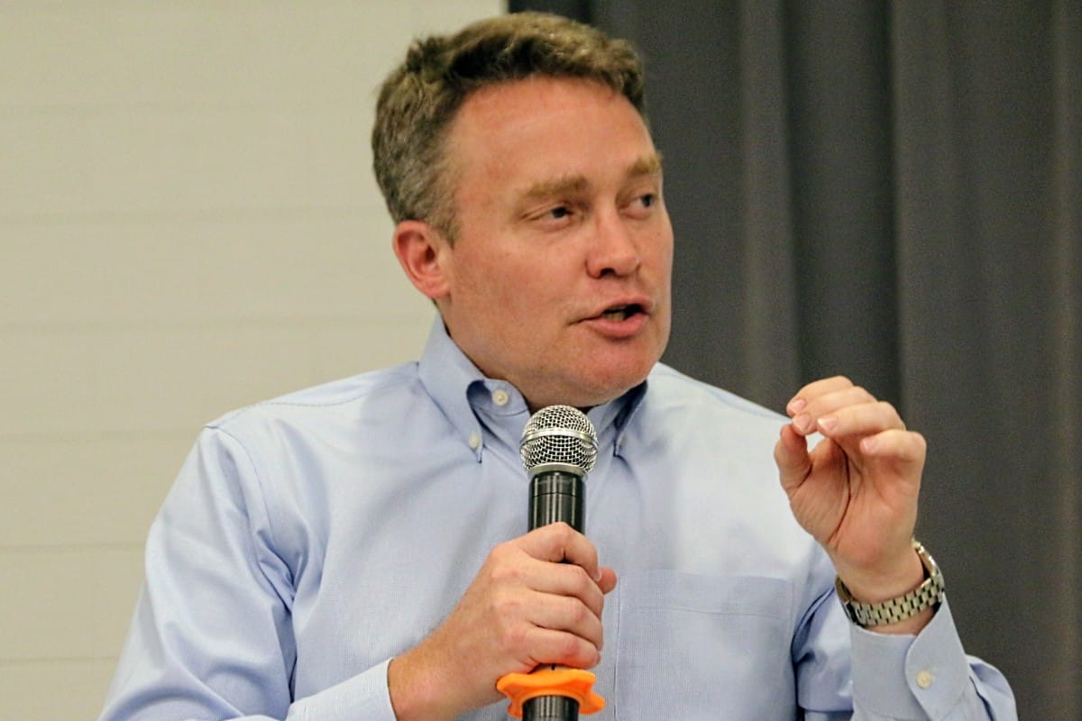 Man in a light blue dress shirt speaks into a handheld microphone, gesturing with his right hand on stage in front of a gray curtain.