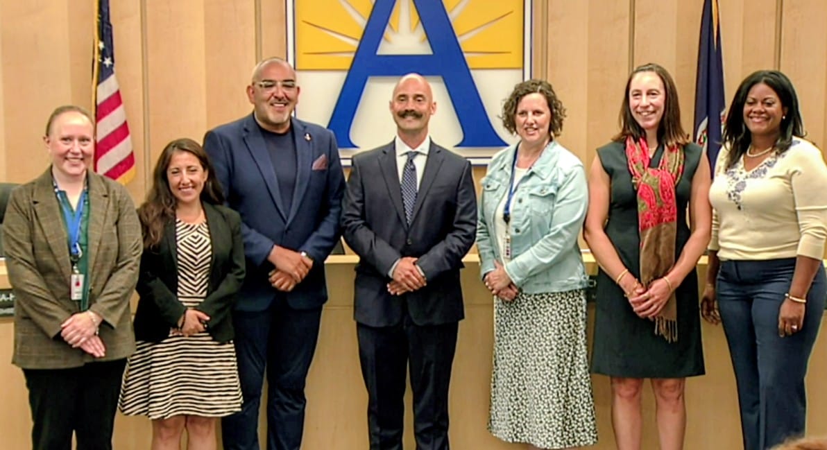 Daniel Bernhardt with School Board members (screenshot via APS) | ARLnow Group of seven professionals in business attire standing side by side in a conference room, in front of a large blue A logo and flags on either side.