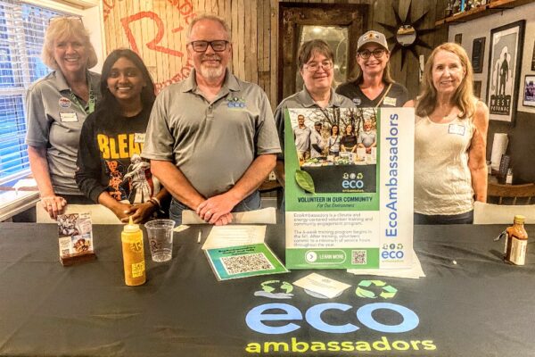Six volunteers at a community table for EcoAmbassadors, with brochures and signage in the background.