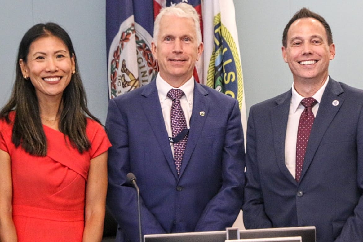 Three adults in business attire pose for a photo at a briefing, with flags in the background (woman in red on the left, two men in blue suits).