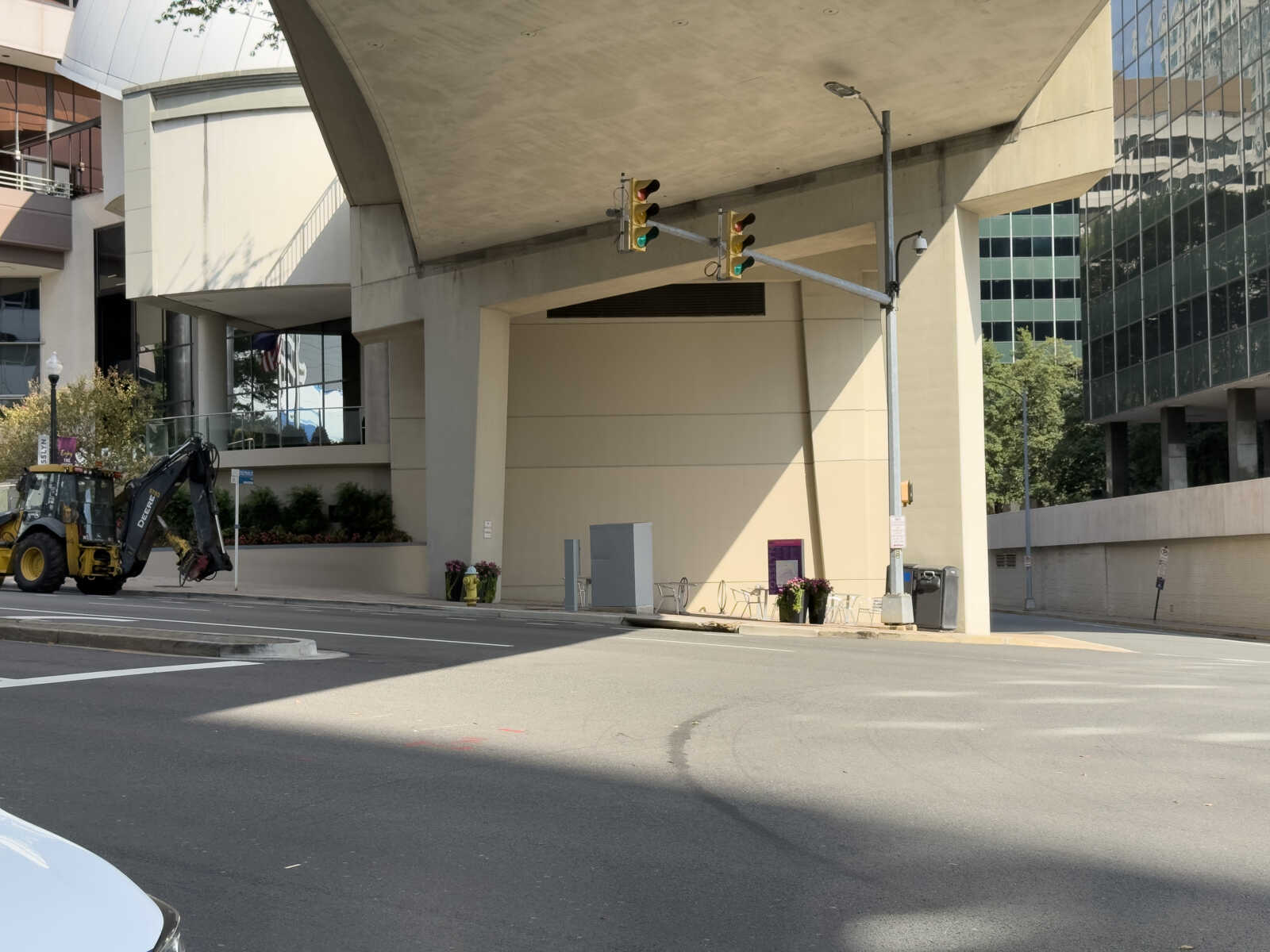 Construction backhoe parked on a city street beside a modern building under a concrete overhang, with traffic lights overhead.