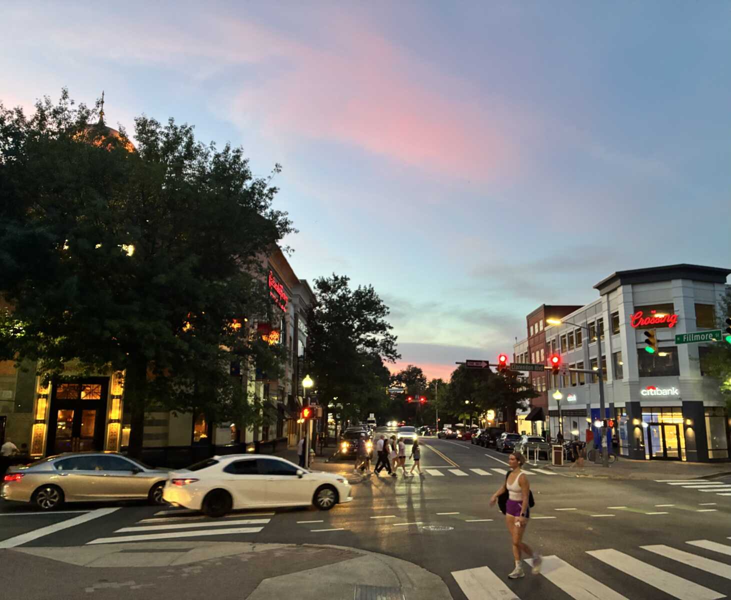 Dusk in Clarendon (courtesy George Brazier) | ARLnow Urban intersection at dusk with pedestrians crossing a crosswalk, cars driving by, and storefronts on a tree-lined street with warm lights.