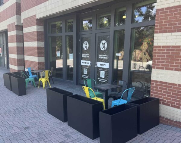 Outdoor cafe seating in front of a brick building, with black rectangular planters and multicolored metal chairs around black tables.