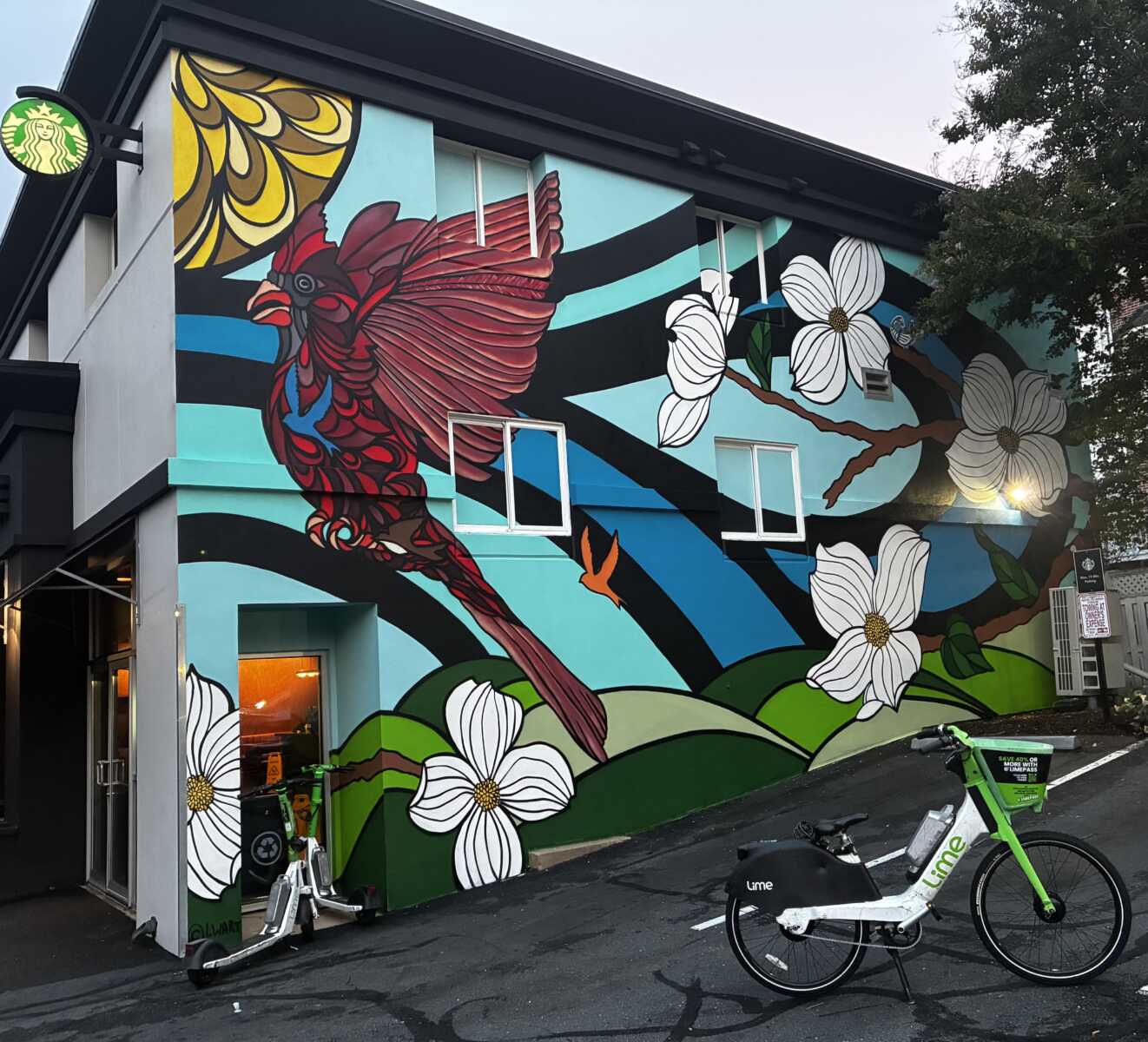 Lime bike parked outside Starbucks in Clarendon (courtesy George Brazier) | ARLnow Colorful mural on a building featuring a red bird, white magnolia blossoms, blue waves, green hills, with a Lime bike and a scooter parked in front.