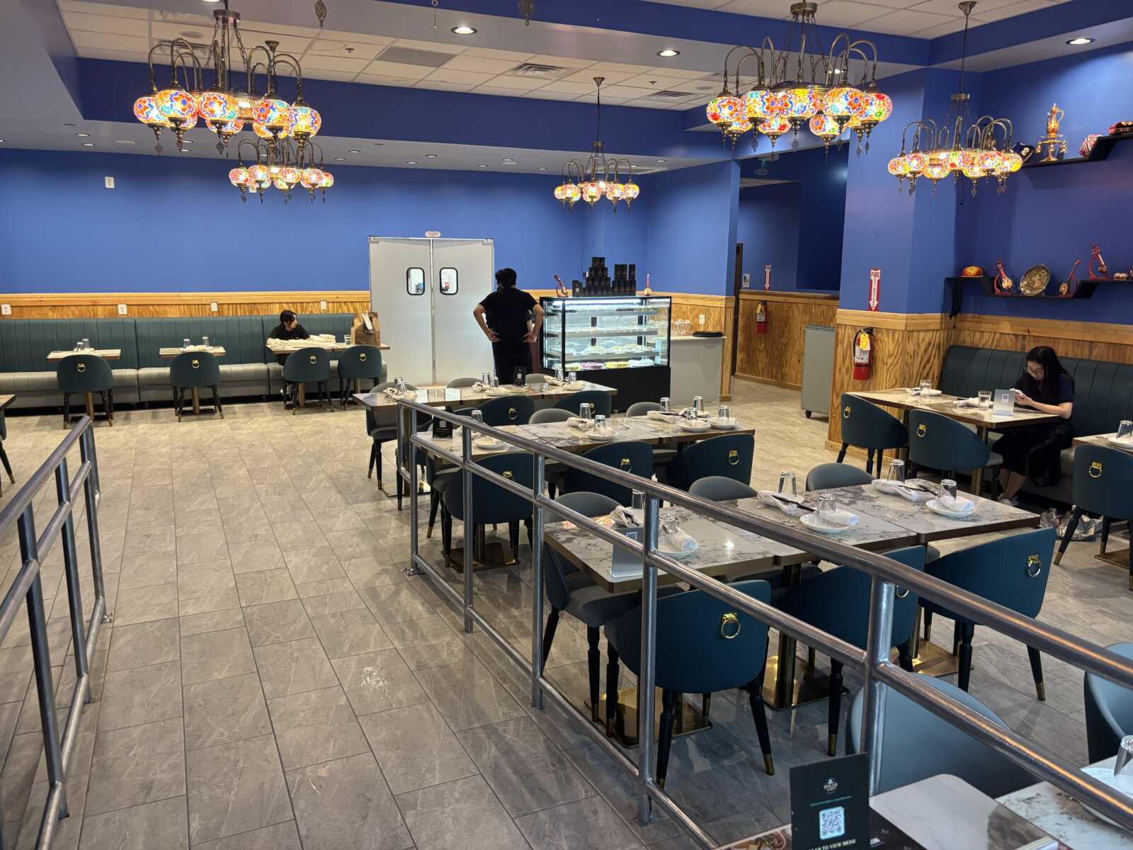 Restaurant dining area with blue walls, teal chairs, and ornate chandeliers; a staff member stands near a pastry display case.