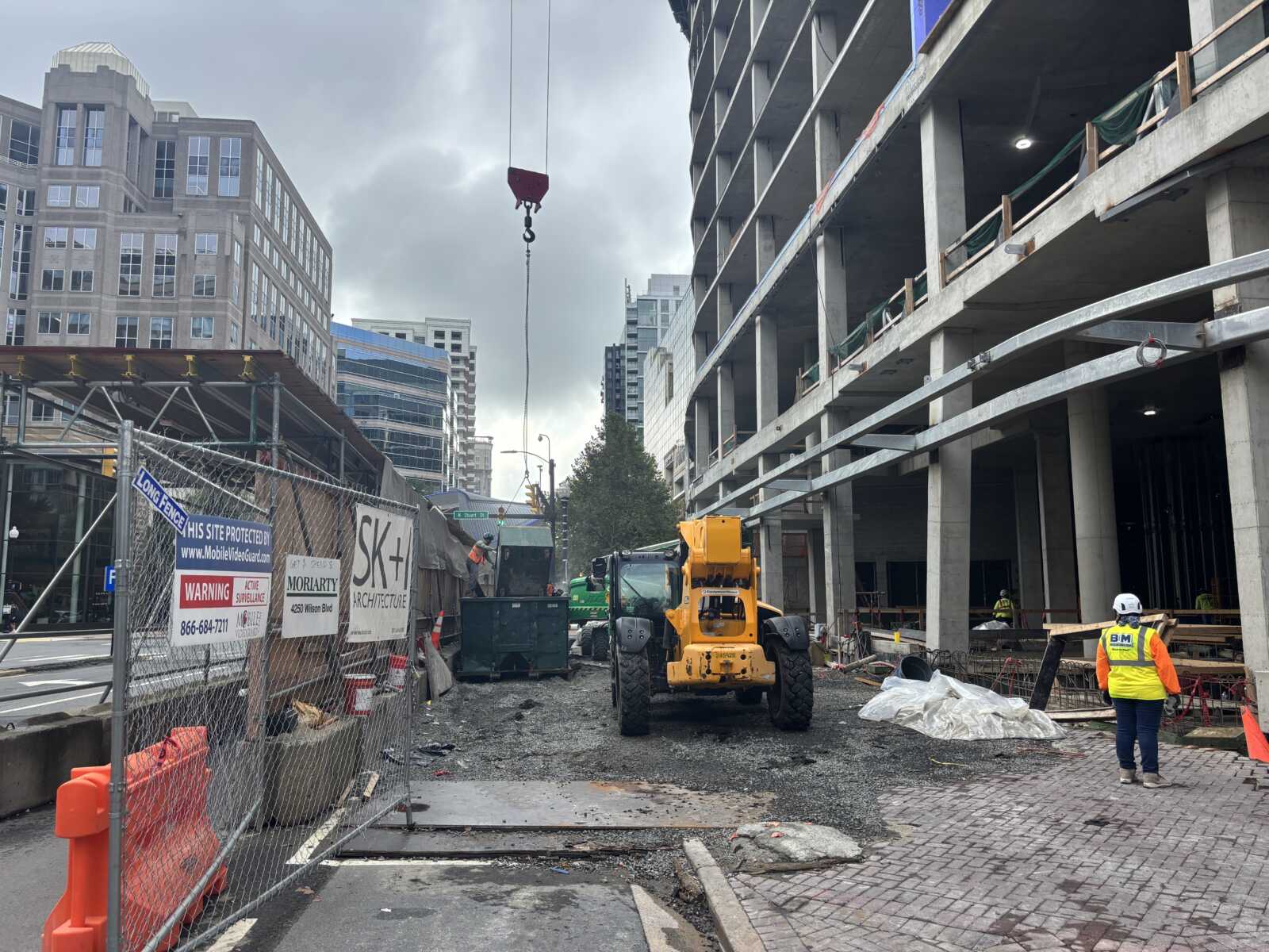 Construction site on a city street with an unfinished multi-story building, yellow loader, and crane hook overhead; workers in safety gear nearby.