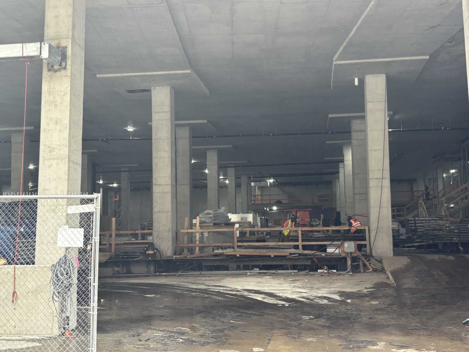 Interior of a large construction site with concrete columns, scaffolding, and workers in high-visibility vests nearby.