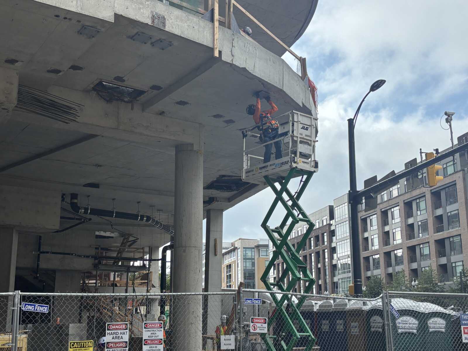 Construction worker in an orange safety vest and hard hat stands on a scissor lift under a concrete overhang, inspecting/welding the beam while a construction site fence with danger signs surrounds the area.
