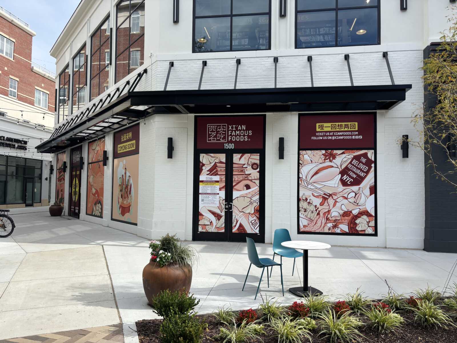 storefront of Xi'an Famous Foods with maroon signage and illustrated window panels; outdoor seating with turquoise chairs and a round table in front.