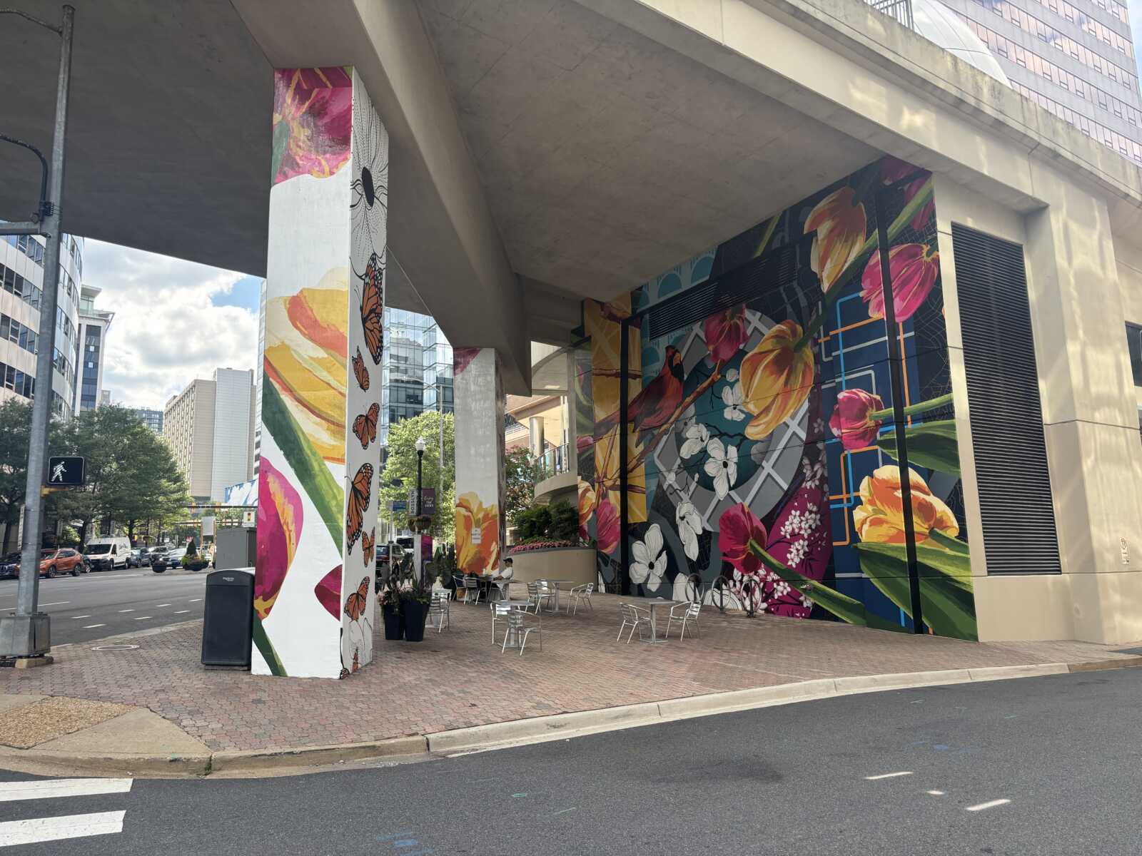 Urban street scene under an overpass with colorful mural-covered pillars and outdoor seating.