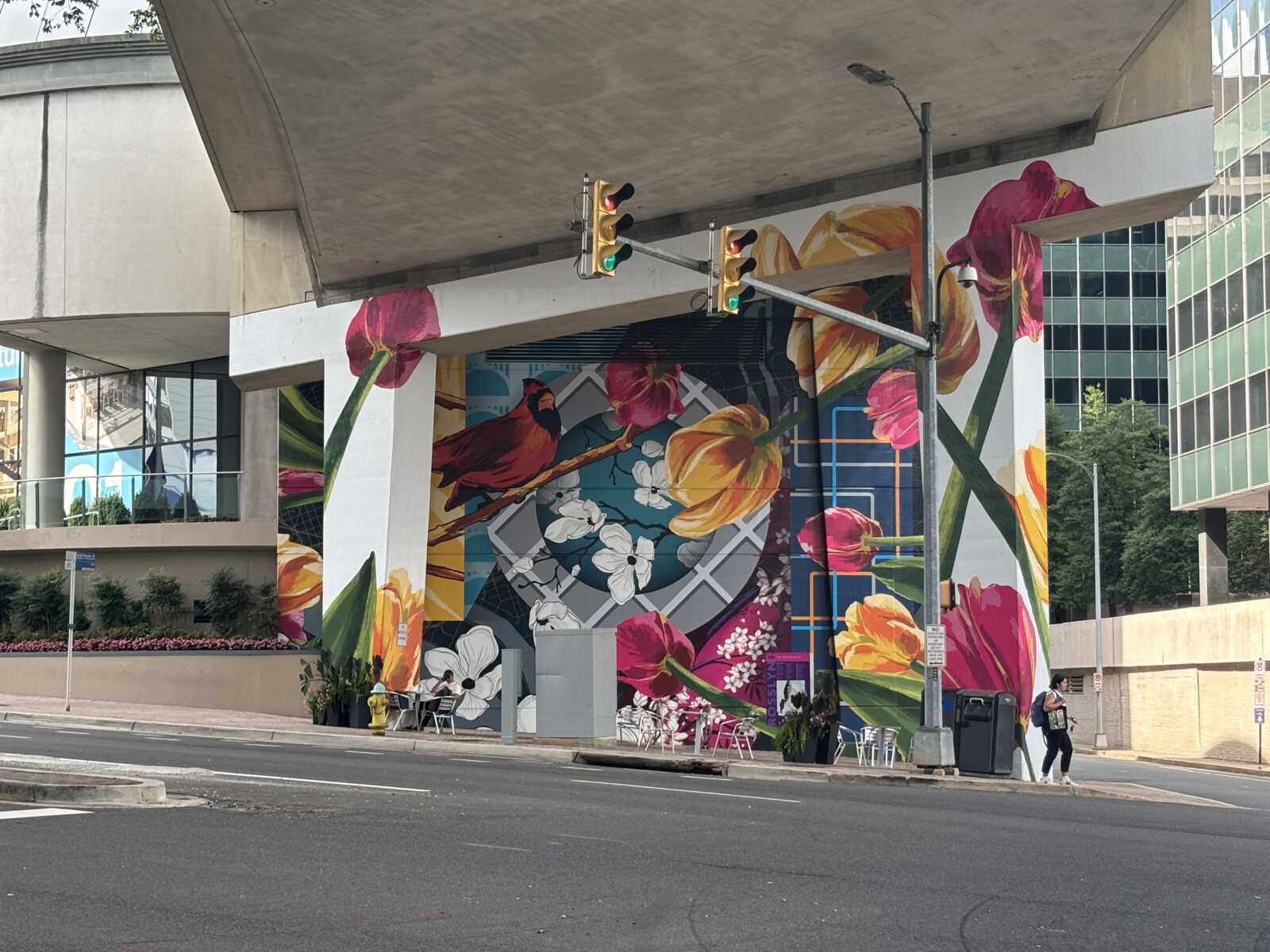 Colorful mural on a building wall featuring large flowers, a red bird, and geometric patterns, under an overhang with a street scene below and traffic lights overhead.