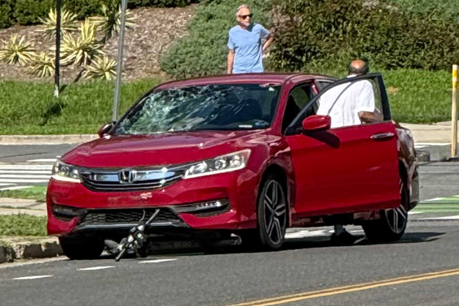 Red Honda sedan with a damaged windshield and front bumper, parked on a street; a man in a blue shirt and sunglasses stands on the roof while another man in a white shirt stands by the open driver’s door. A bike or tripod-like object lies under the car’s front bumper.