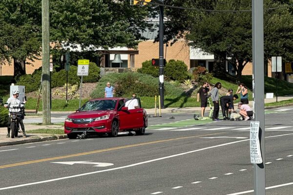 Red sedan stopped in the crosswalk with its driver door open, while a group of people attends to someone on the pavement near a parked scooter on the curb and a cyclist waits nearby on the left.