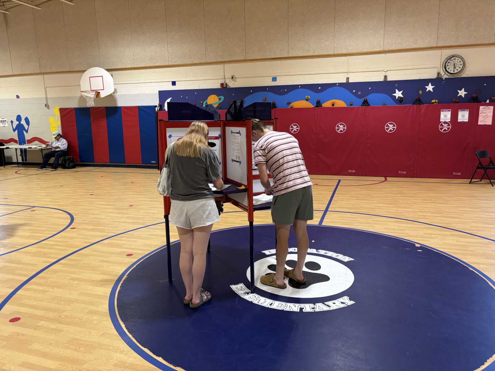 People vote at a red ballot booth set up in a school gym, with a colorful padded wall and wall mural in the background.
