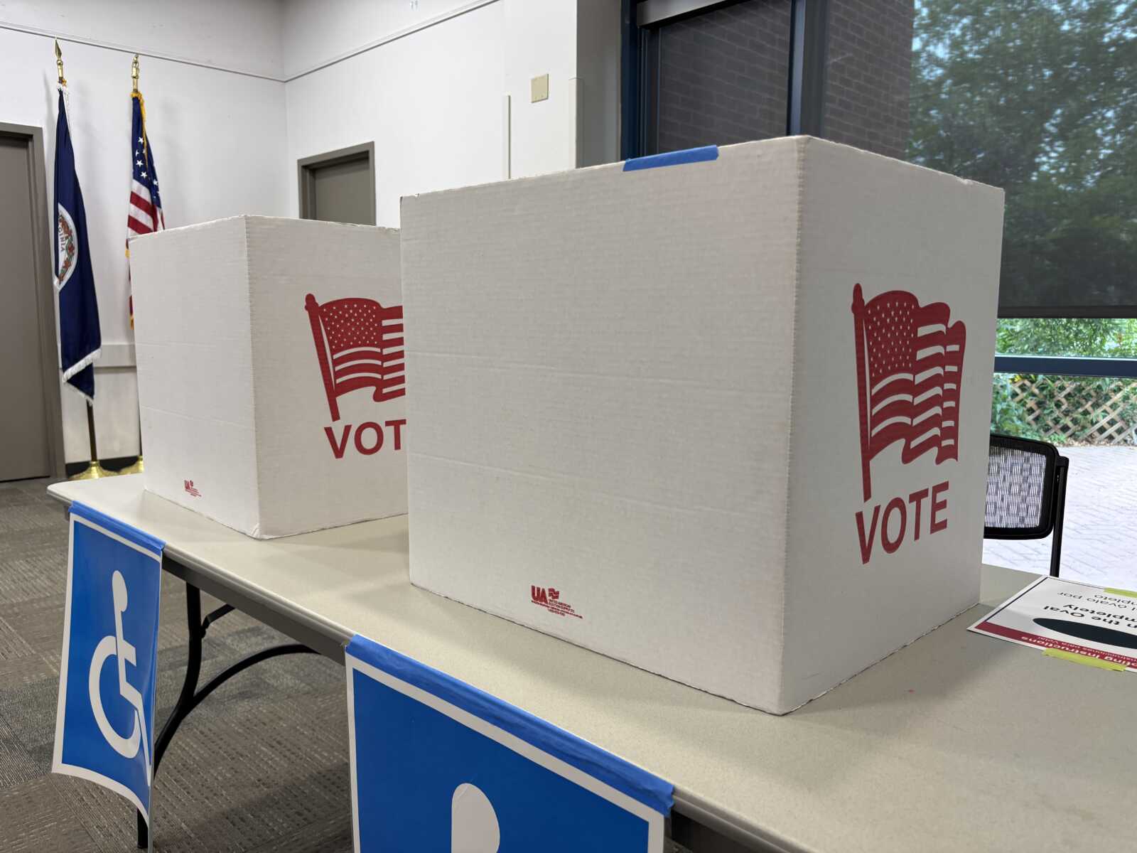 Two white cardboard voting booths labeled VOTE sit on a table in a room with US flags in the corner and blue accessibility banners along the front.