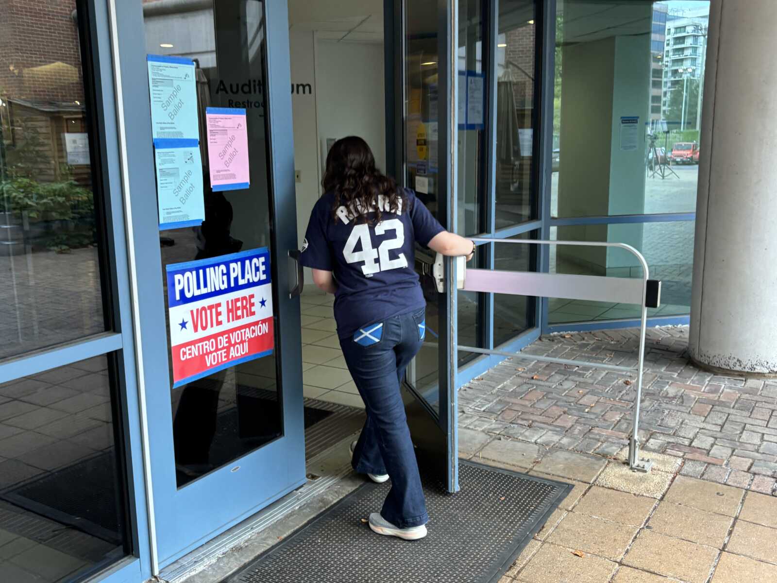 Voter in a navy shirt with the number 42 enters a polling place through glass doors with a blue-and-red sign that says 'POLLING PLACE VOTE HERE'.