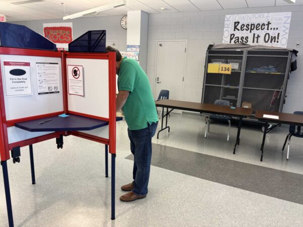 Voter standing at a red polling booth placing a ballot inside a slot in a bright room with tables in the background, indicating a polling station.