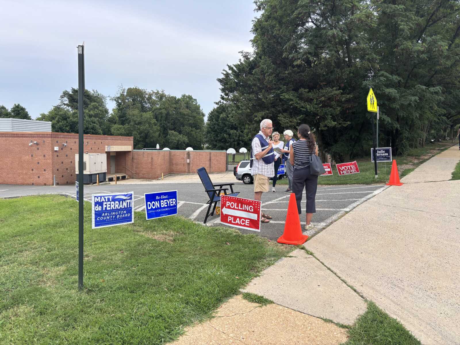 Voters and volunteers stand by a polling place with blue and red campaign signs and orange cones nearby.